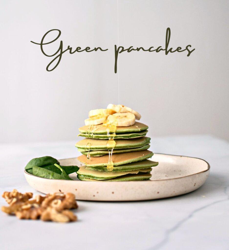Stack of fluffy green spinach pancakes topped with sliced bananas and drizzled with honey, served on a speckled plate with fresh spinach leaves and walnuts.