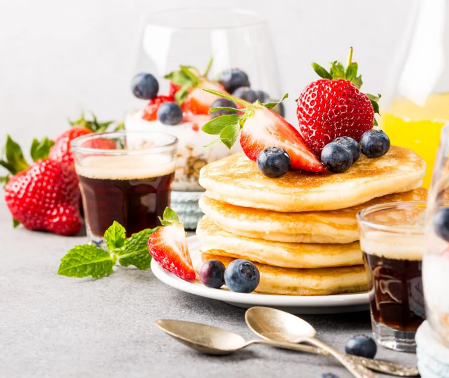 Golden pancake stack topped with fresh strawberries and blueberries, served on a bright breakfast table with black coffee, orange juice, and yogurt parfaits.